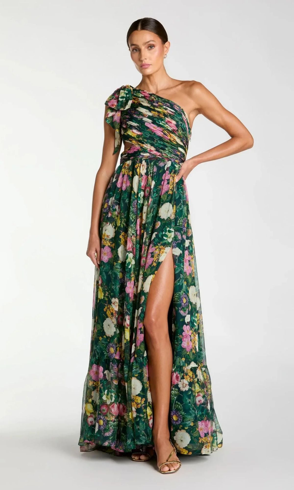 A woman poses against a plain background in the Mac Duggal 68553 long formal dress, a deep green multi floral chiffon one-shoulder evening gown with a side slit and bow detail on the shoulder.